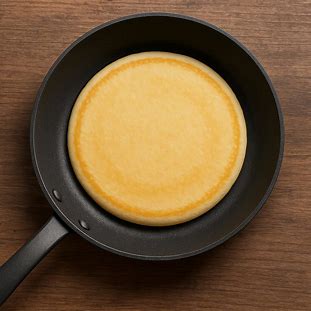 Imagem de uma frigideira com massa de panqueca cozida, mostrando textura lisa e cor dourada clara.