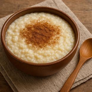 Tigela de mingau de mandioca cremoso com canela em pó e colher de madeira ao lado.