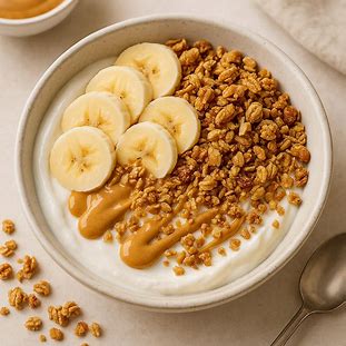 Bowl de Iogurte Grego, Granola e Pasta de Amendoim. Foto de uma tigela de iogurte branco cremoso, coberto por fatias de banana, granola e um fio de pasta de amendoim.