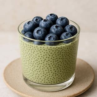 Pudim de Chia com Matcha. Foto de uma sobremesa ou café da manhã em um copo de vidro. O pudim tem uma cor verde pálida (do matcha) e é coberto por mirtilos frescos.