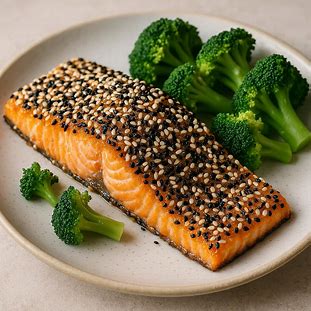 Filé de Salmão Assado com Crosta de Gergelim. Peixe com cor alaranjada, coberto por uma camada crocante de gergelim branco e preto, servido ao lado de brócolis cozidos no vapor.