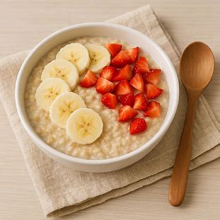 Texto alternativo para imagem: Tigela branca contendo mingau de aveia cremoso e quente, decorado com fatias de banana e pedaços de morango frescos, ao lado de uma colher de madeira sobre um pano de cozinha bege, em uma superfície de madeira clara.
